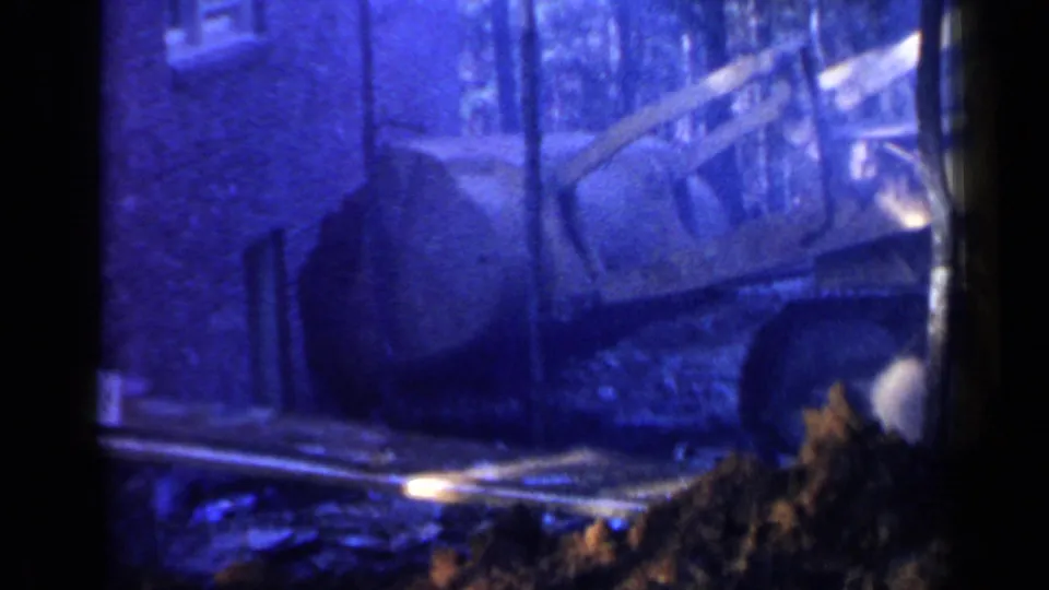 The excavator bucket and arm sit low in the frame amid trees, dirt, and heavy blue color shift from the 8mm scan.