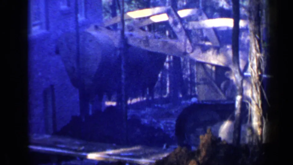 1957 Birmingham home-movie frame with a bulldozer bucket near piled dirt, framed by trees and a nearby wall.