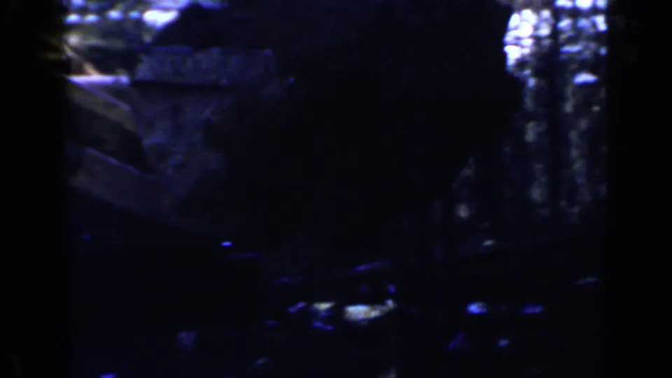 Low-light 1957 Birmingham 8mm frame showing wooded ground, rocks, and a shadowed construction vehicle shape near the trees.