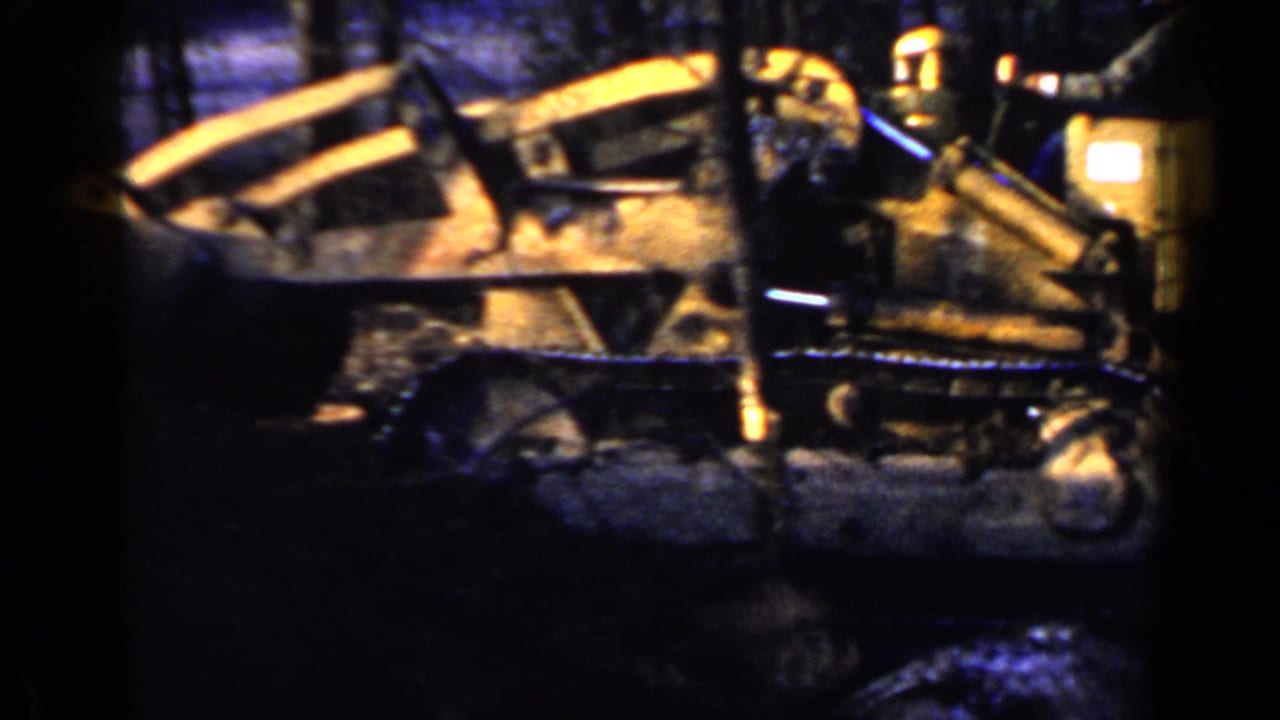 Yellow tracked tractor seen from the side among trees on dark red soil.