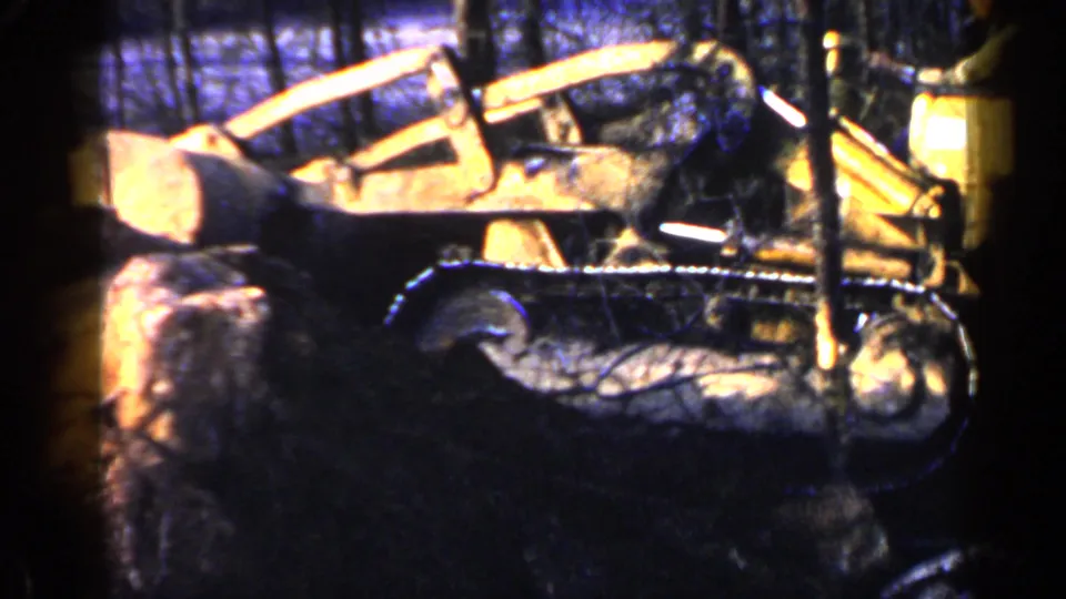 A yellow tracked tractor appears in profile in a wooded Birmingham, Alabama setting, partly blocked by a foreground tree.
