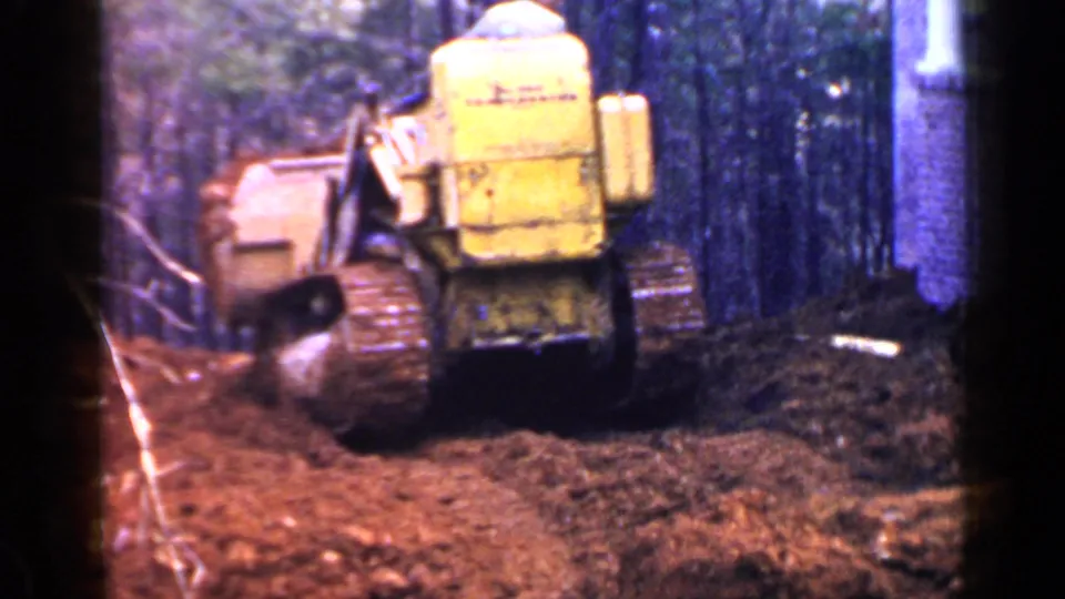 Rear view of the yellow tracked tractor on churned red earth, with trees and a brick structure edge nearby.