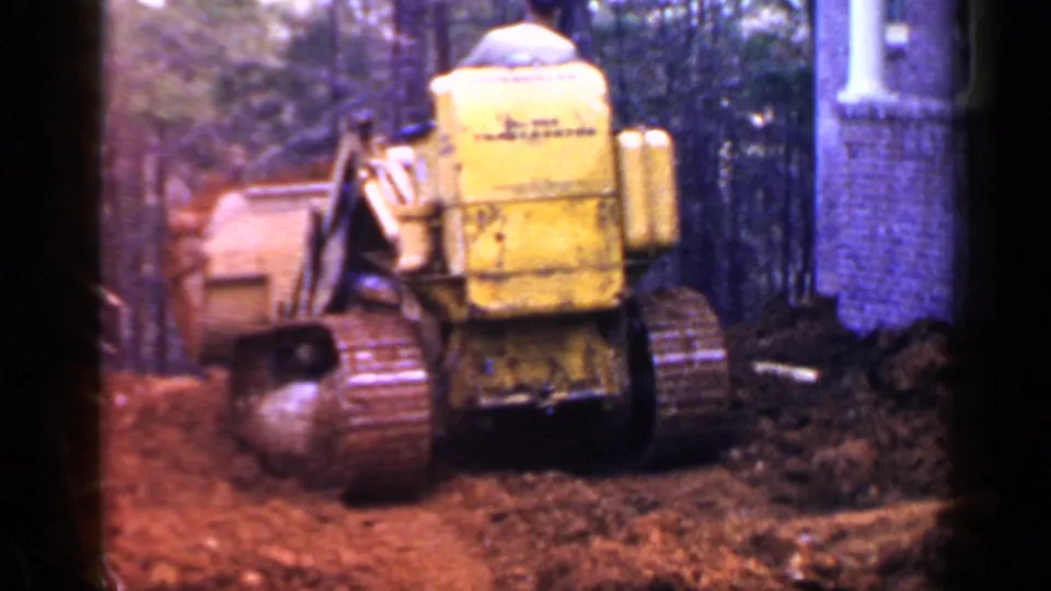 1957 home-movie frame showing the rear of a yellow tracked tractor on red earth beside trees and a brick structure.