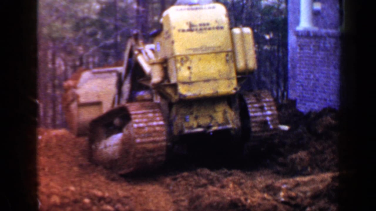Yellow tracked loader seen from behind moving dirt near a brick building.