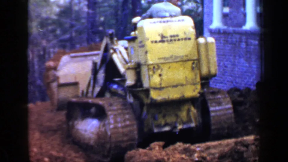 The yellow construction machine appears angled across churned soil, with brickwork, a window, and white columns visible in the background.