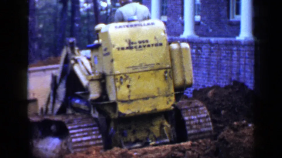 A close rear view shows the yellow loader, its tracks, bucket edge, dark soil, and the brick building behind it in 1950s Birmingham footage.