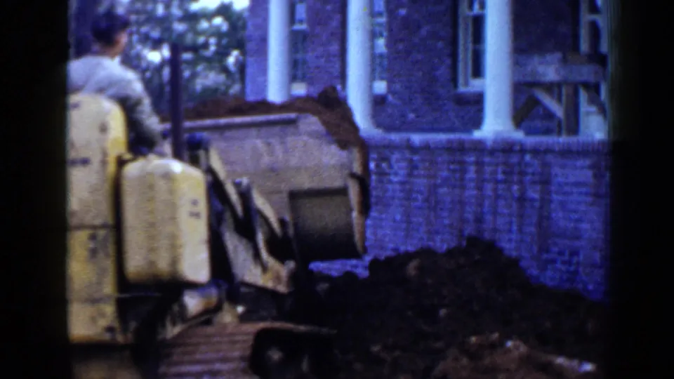 A man in a green shirt operates the yellow tracked loader near churned soil and a brick building with white columns in Birmingham, 1957.