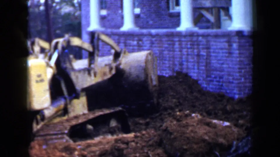 A yellow tracked bulldozer is seen from the side as it works the front yard outside the brick house.