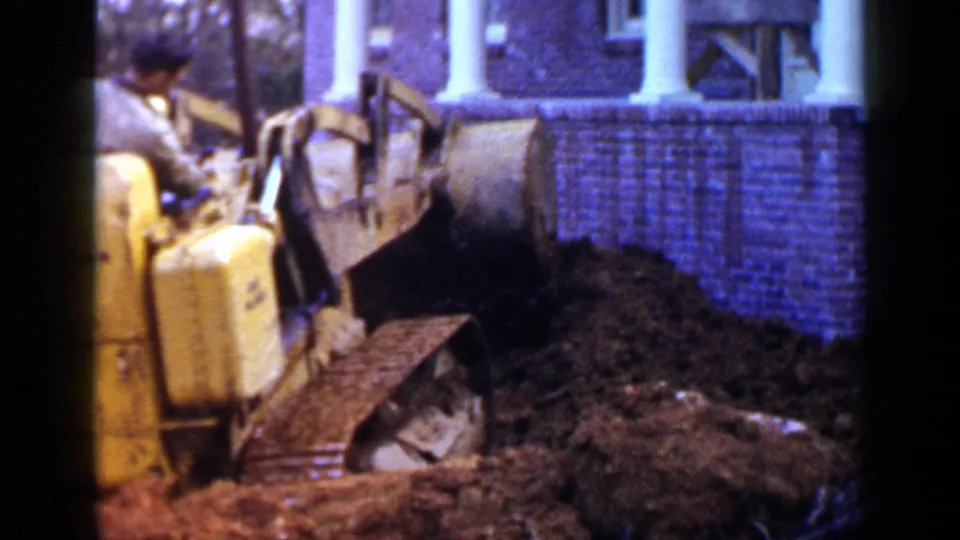 A man operates the yellow bulldozer in the front yard of a brick Birmingham house, with the bucket in loose dirt.