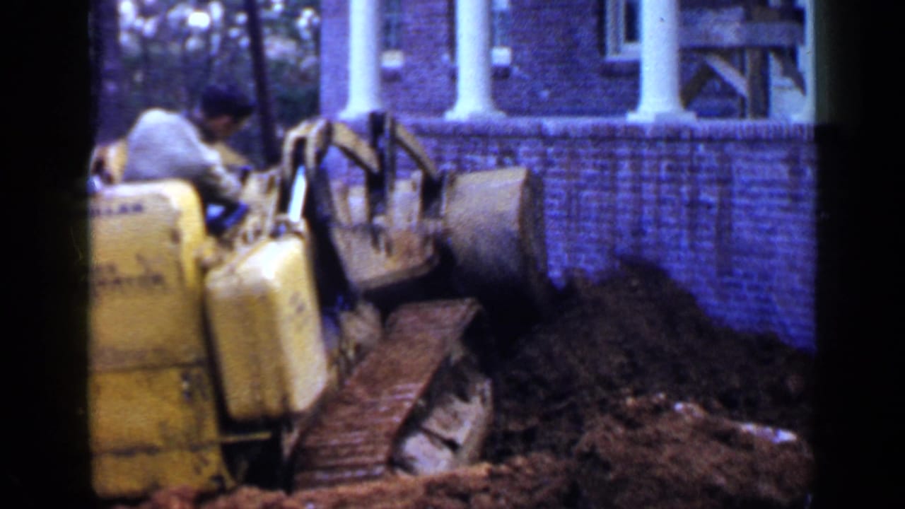 Yellow bulldozer beside a brick building pushes dirt near trees.