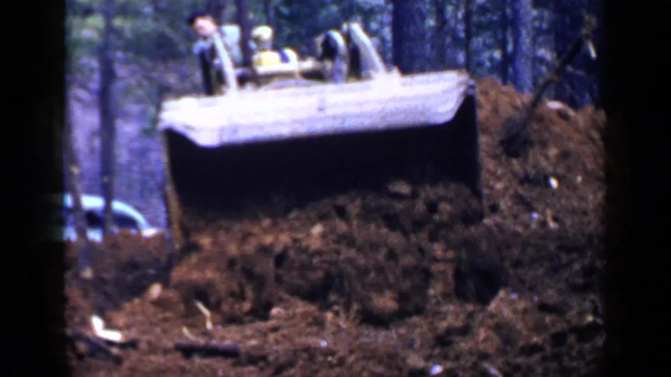 A yellow bulldozer in 1957 Birmingham footage pushes loose soil through a wooded area, with the operator visible above the blade.