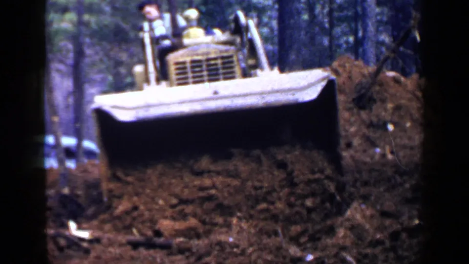 The front of a yellow bulldozer fills the frame as it moves soil in a wooded Birmingham, Alabama setting in 1957 footage.