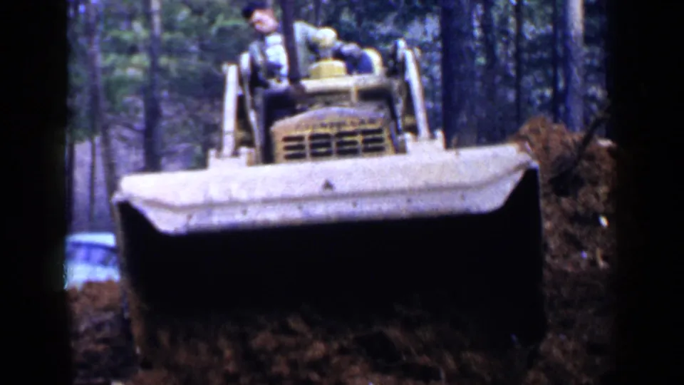 A yellow bulldozer approaches in wooded 1957 Birmingham home-movie footage, pushing a pile of soil with the operator visible above.