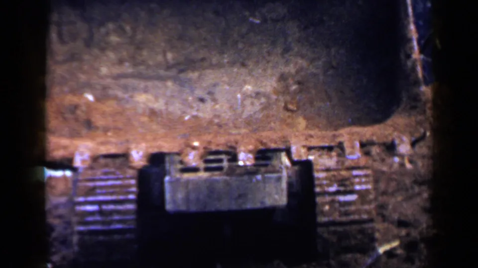 Close 8mm view of the bulldozer undercarriage and wide blade pushing through dark soil at the wooded work site.