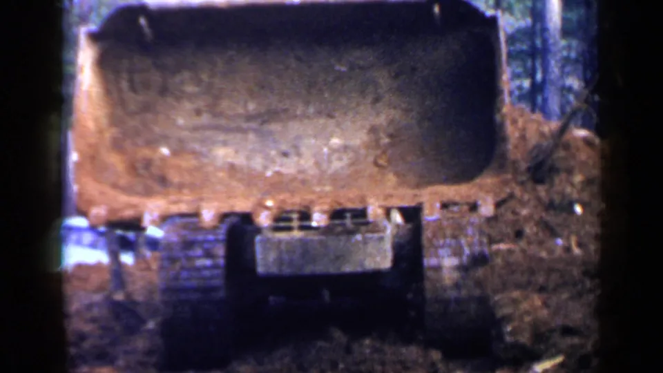 Close 1957 Birmingham-area frame of a crawler bulldozer blade and tracks moving earth in a wooded construction setting.