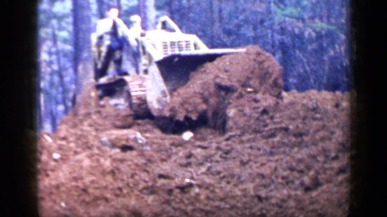 Tracked bulldozer pushes a mound of dirt in a wooded clearing with two workers aboard.