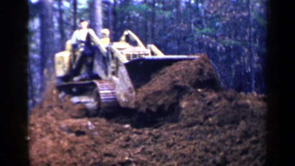 1957 Birmingham home-movie frame of a bulldozer carrying soil across churned ground in a wooded area with two workers aboard.