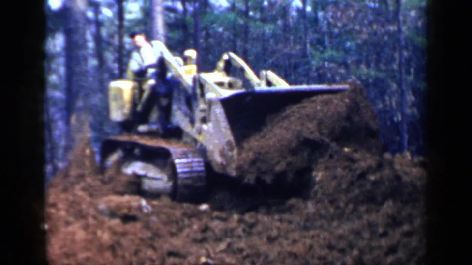 1957 Birmingham 8mm frame showing the side of a yellow bulldozer pushing soil, with two workers visible against dense trees.