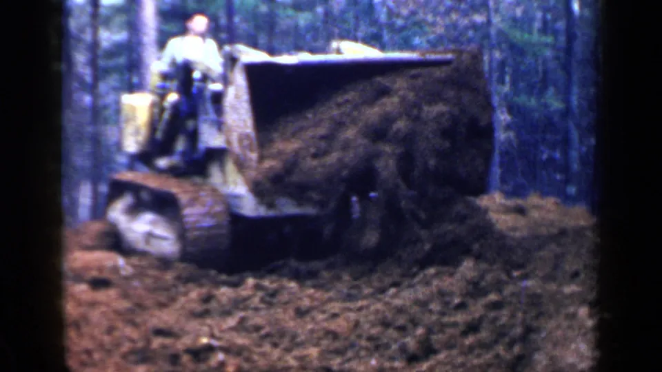 1957 Birmingham home-movie frame of a tracked bulldozer with a raised blade full of soil in a forest clearing.