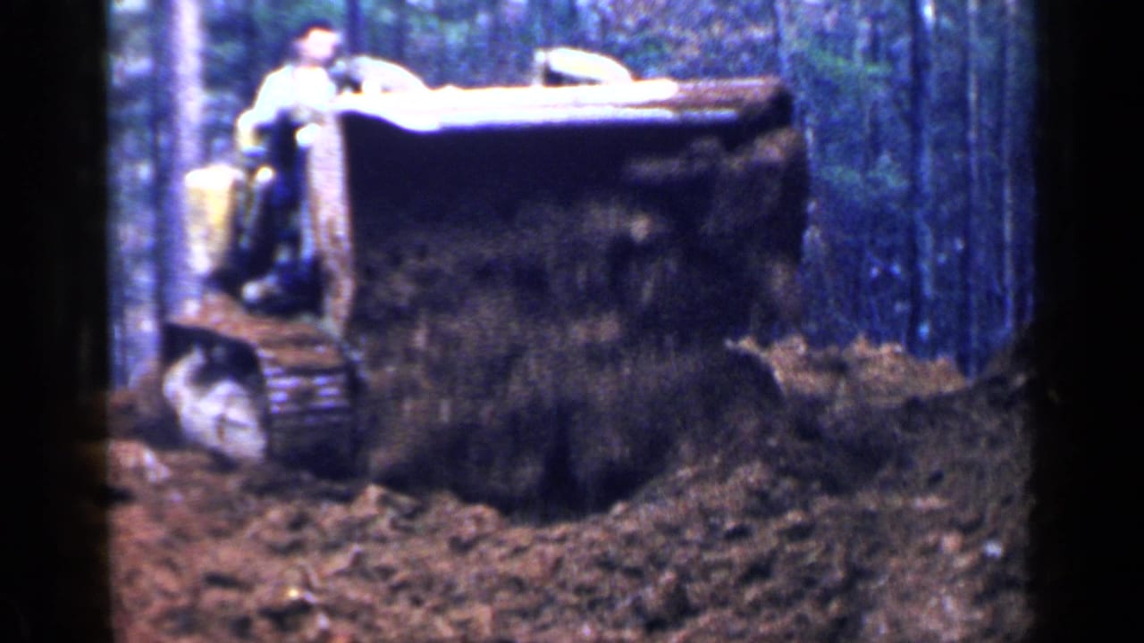 Yellow tracked tractor with front bucket pushing dark soil in a wooded area.