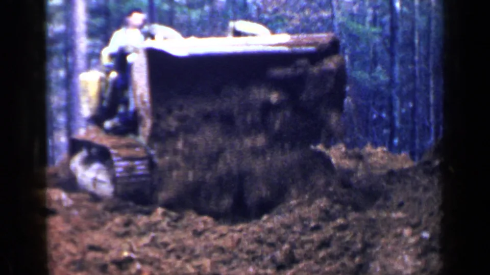 1957 Birmingham home-movie frame showing a man on a yellow tracked tractor moving earth among trees.