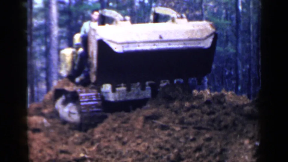 The yellow tractor raises its bucket over a growing dirt pile in wooded Birmingham-area footage from 1957.