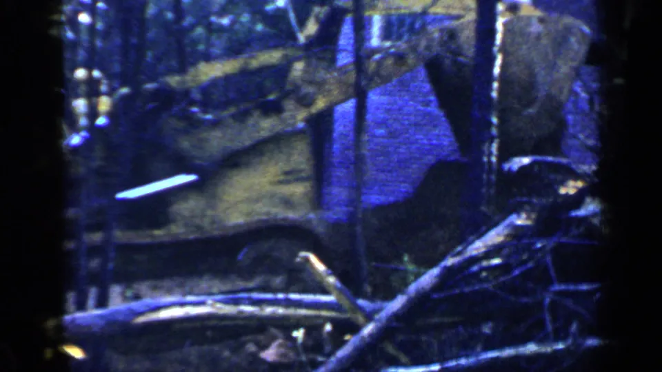 A closer 1957 Birmingham home-movie view shows the tractor near felled wood and trees, with the operator partly visible.