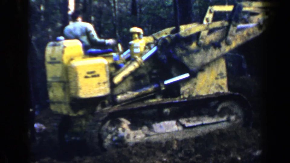 A worker in a light shirt and cap operates a yellow tracked bulldozer in a wooded Birmingham setting in 1957 footage.
