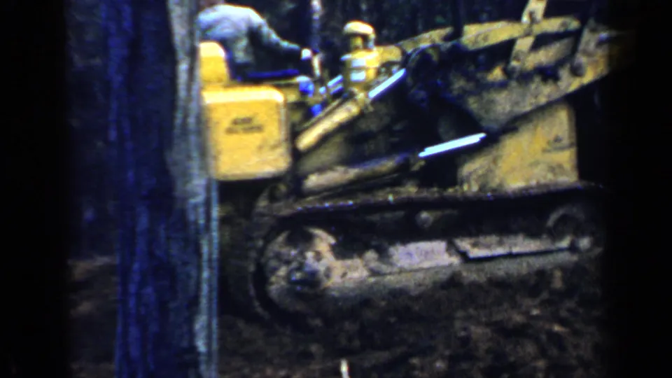 1957 Birmingham home-movie frame showing a yellow tracked bulldozer moving through a wooded path with the operator partly seen.