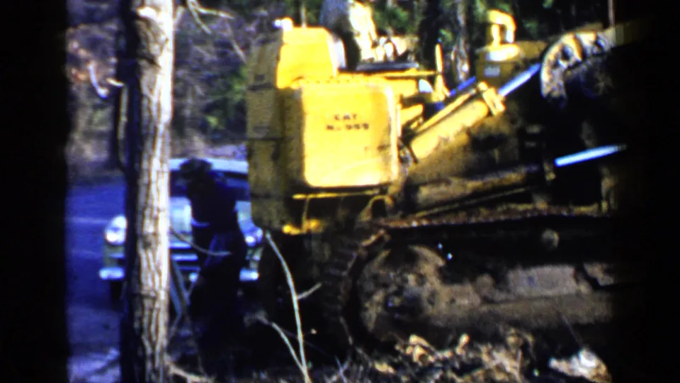 Yellow bulldozer passes through a wooded area while part of an older car appears in the background of the Birmingham home movie.