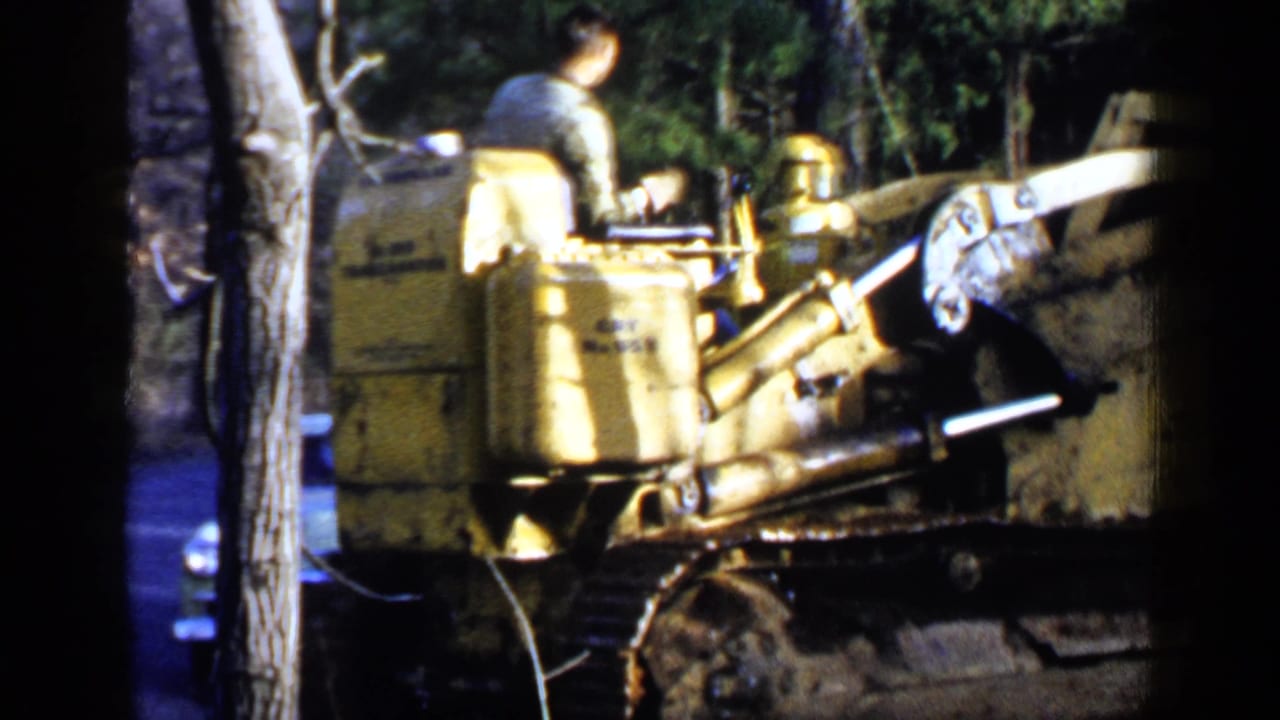 Man operating a yellow tracked bulldozer among trees with another person and a car partly visible.
