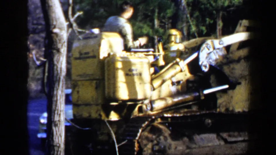 1957 Birmingham-area 8mm frame showing a man on a yellow tracked bulldozer in woods, with another person and a vintage car partly visible.