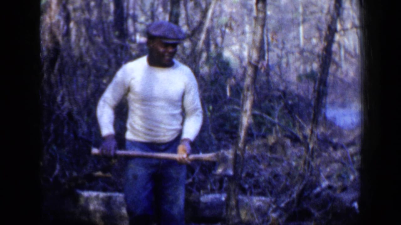 Man in a white sweater and cap stands in brush holding an axe or long-handled tool.