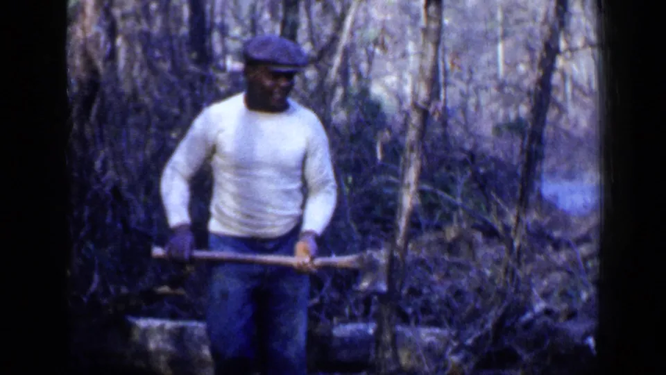 1957 Birmingham-area 8mm frame showing an adult man in a white sweater and cap holding an axe in dense woods.