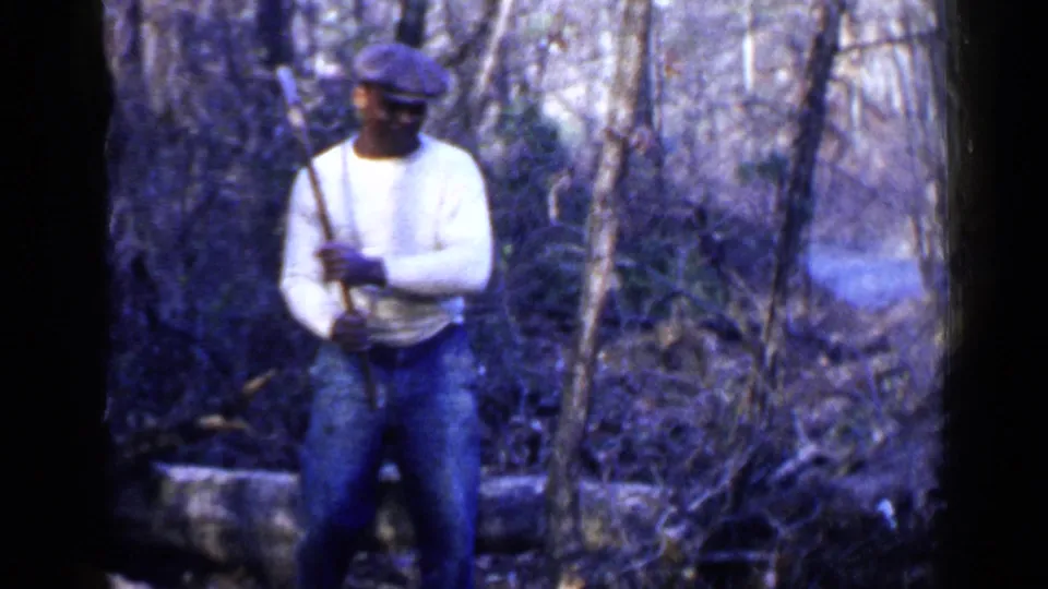1957 Birmingham-area 8mm frame showing a man holding a tool upright in a wooded area with brush and fallen wood around him.