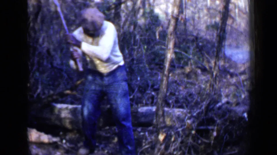 1957 Birmingham-area 8mm frame showing a man in a white sweater bending with an axe near fallen wood in dense brush.