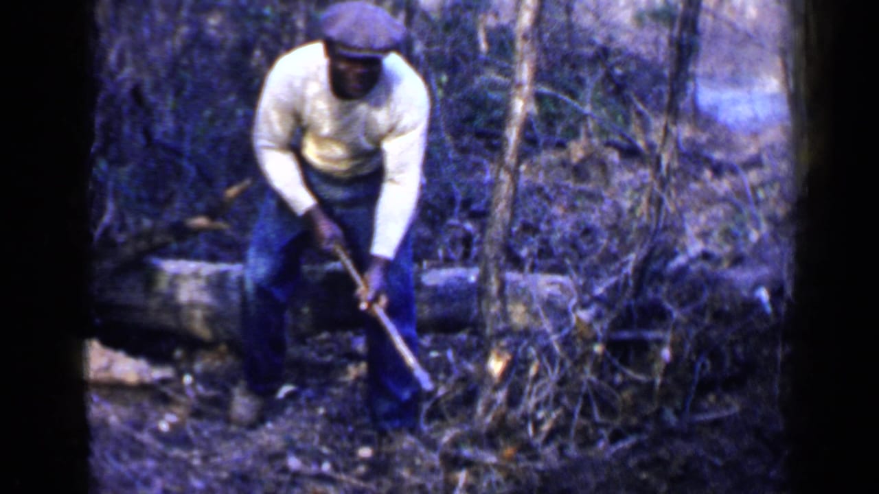 In 1957 Birmingham home-movie footage, a man in a white sweater and flat cap bends over a long-handled tool in a wooded area.