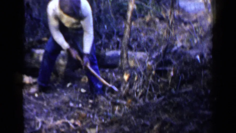 A man in mid-century outdoor clothing bends into the work, using a long-handled tool near brush and a fallen log in Birmingham.