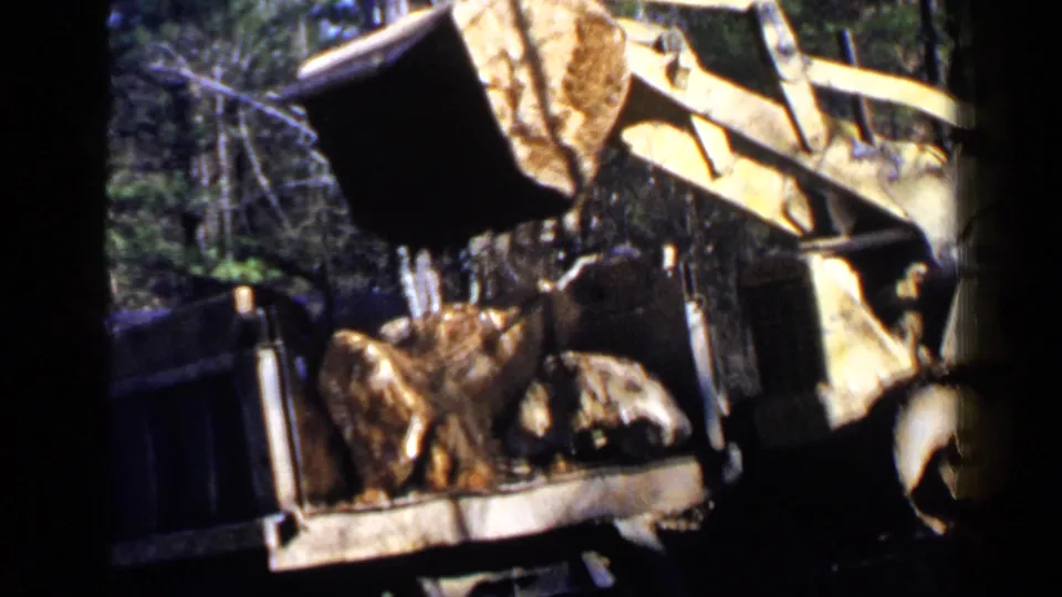 A yellow front-end loader tips a bucket of rocks into a trailer or truck bed, filmed on grainy 8mm home-movie stock.