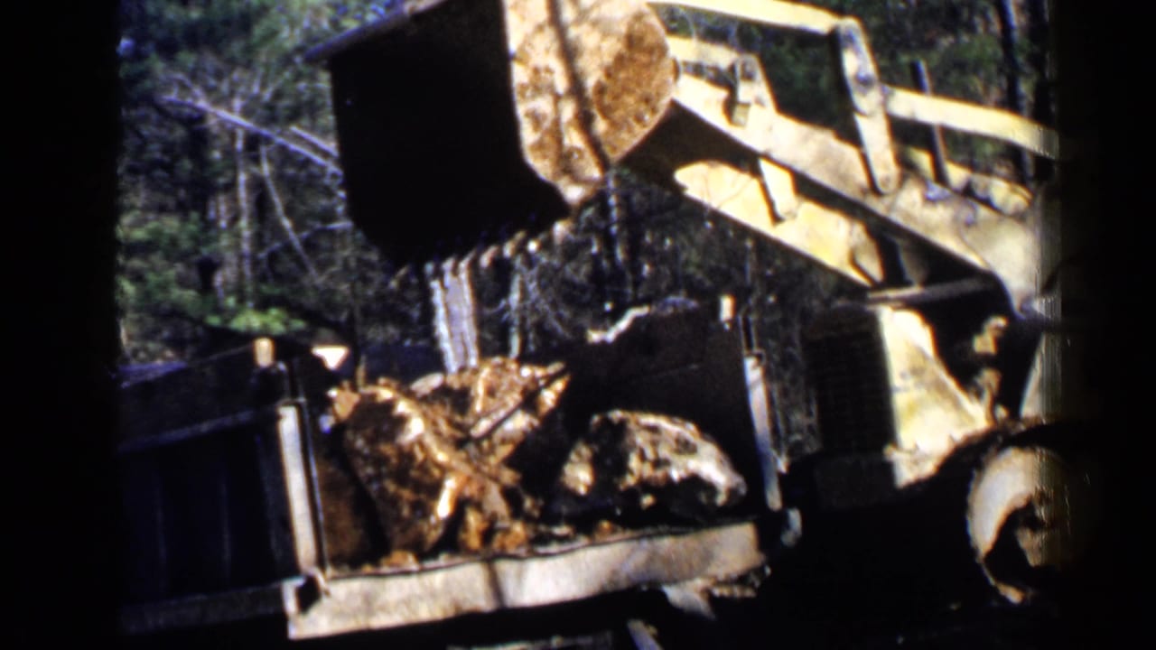 Front-end loader raises a bucket of rocks beside a dump truck in a wooded work area.