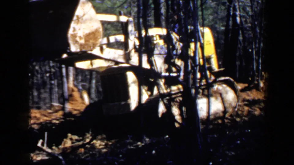 A 1957 8mm view of the loader bucket lifted above a wooded work area, with truck parts and debris visible below.