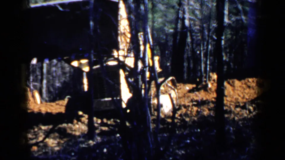 Loader and truck remain in the wooded Birmingham-area site, with a partly visible worker or bystander near the equipment.