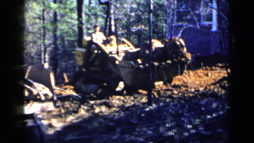 1957 Birmingham frame showing a bulldozer operator working amid trees and churned soil near a residential building.