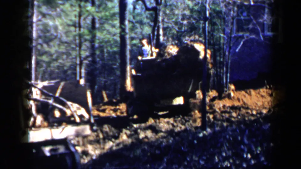 1957 Birmingham-area 8mm frame showing a bulldozer in a wooded clearing, with earth, debris, and a partial building wall visible.