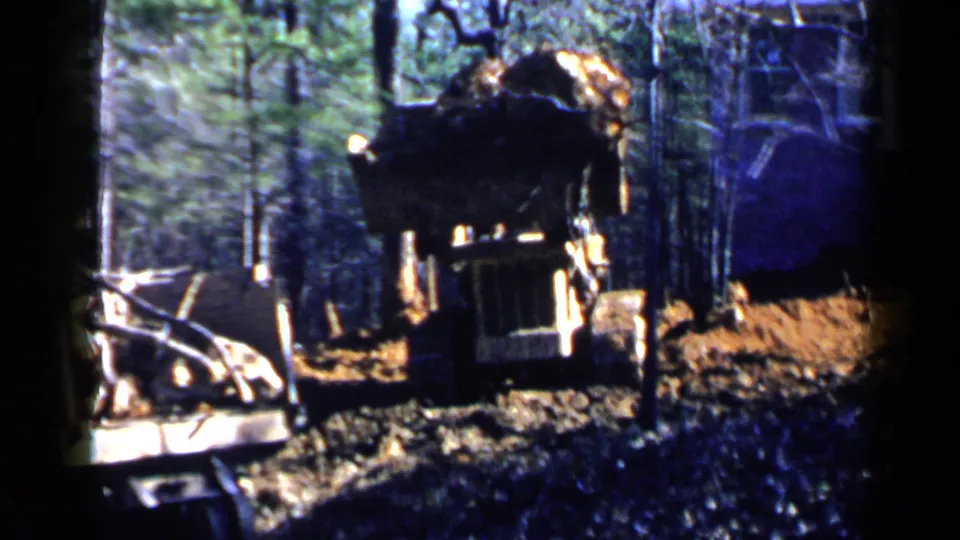1957 Birmingham-area home-movie frame with a front view of the bulldozer in a wooded clearing beside debris and a nearby structure.