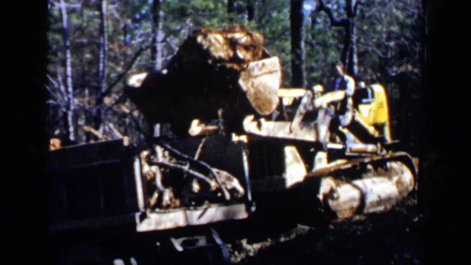A yellow front-end loader lifts roots or debris toward a dump truck in 1957 Birmingham home-movie footage.