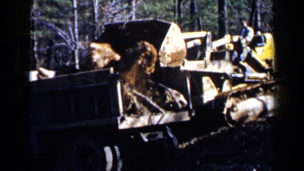Yellow earthmoving machine loads large rocks into a dark dump truck in a wooded work area.
