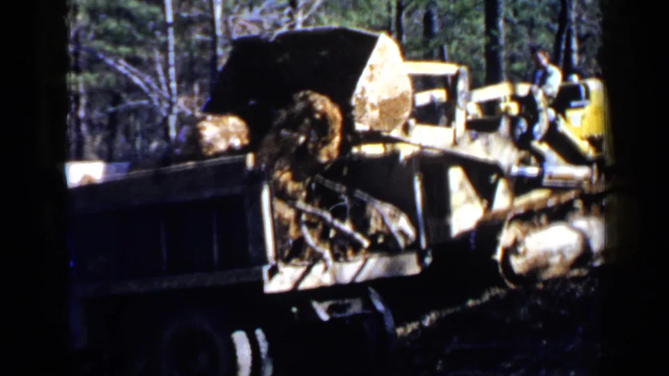 Yellow heavy equipment works beside a dark dump truck in wooded Birmingham-area footage from 1957, with rocks visible in the bed.