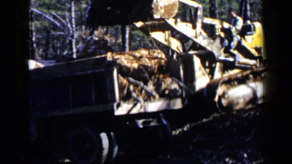 1957 8mm frame of a yellow bulldozer or loader tipping rocks and debris into a dark dump truck in a forested work area.