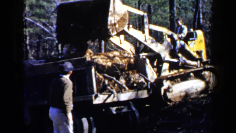 An adult man observes a yellow bulldozer or loader beside a dark dump truck in 1957 Birmingham-area wooded construction footage.